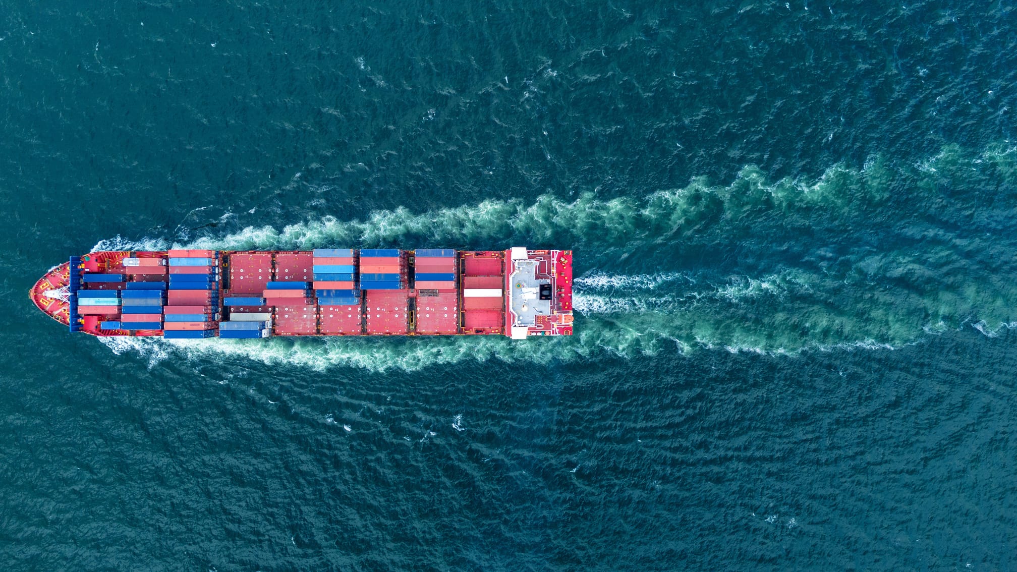 Luftaufnahme eines großen Containerschiffs, das beladen mit Frachtcontainern durch das tiefblaue Meer fährt.