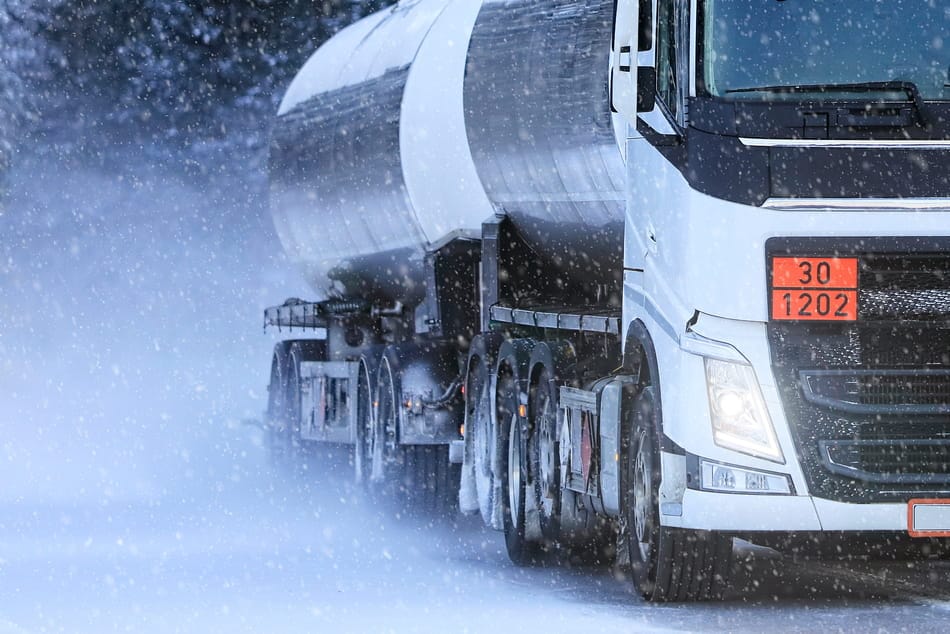 Ein weißer Tank-LKW mit orangefarbener ADR-Warntafel fährt während eines Schneesturms auf verschneiter Straße.