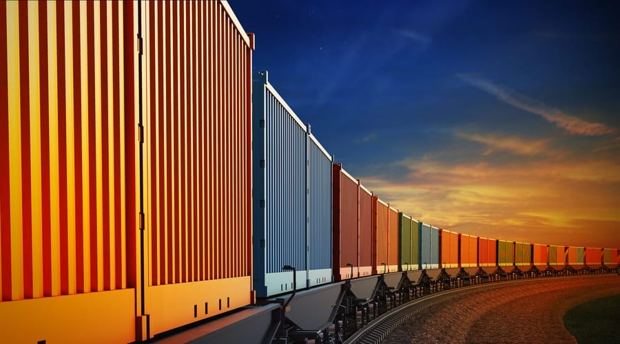 Ein langer Güterzug geladen mit bunten Frachtcontainern fährt auf Schienen in einer Kurve bei einem stimmungsvollen Sonnenuntergang.
